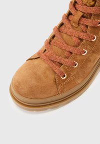 Botín de ante marrón con cordones color óxido y suela de goma texturizada, ubicado sobre un fondo blanco plano.