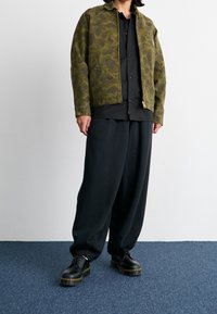 Person som bär en olivgrön kamouflagejacka över en svart knäppskjorta, lösa svarta byxor och svarta läderskor som står på en blå matta.