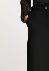 Pantalons noirs ajustés en tissu lisse, avec une poche avant et un bouton rond en laiton à la taille.