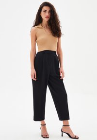 Top a costine beige con scollo a V e colletto. Pantaloni neri a vita alta con pieghe. Tacchi neri con cinturini. Sfondo bianco puro.
