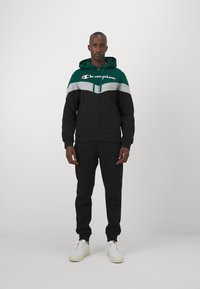 Sudadera con cremallera negra con acentos en verde y gris, que presenta el logo de "Champion". Combinada con pantalones negros a juego y zapatillas blancas.