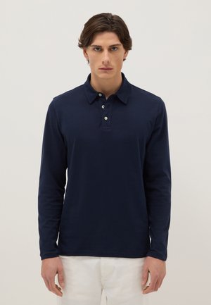 Jeune homme aux cheveux bruns portant un polo à manches longues bleu marine et un pantalon blanc, debout devant un fond clair uni.
