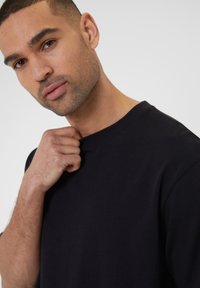 Sort bomulds T-shirt med rund halsudskæring. Modellens hånd griber om kraven og viser stoffets glatte tekstur.