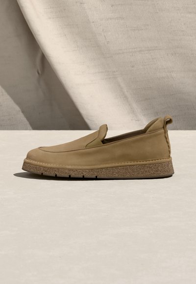 Παπούτσι τύπου slip-on σε ταμπά χρώμα με υφή στη λαστιχένια σόλα και θηλιά στο πίσω μέρος, τοποθετημένο πάνω σε ουδέτερο υφασμάτινο φόντο.