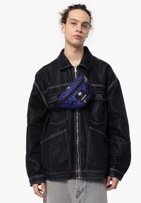 Giacca di jeans nera con cuciture bianche e ampie tasche, con una borsa a tracolla blu a fantasia indossata sopra il torace.