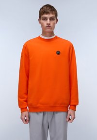 Felpa arancione con colletto rotondo, maniche lunghe e un piccolo logo nero sul petto. Tessuto morbido, abbinata a pantaloni grigio chiaro.