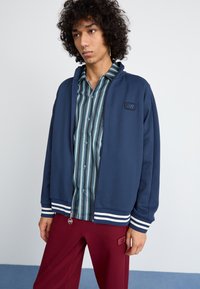 Navy zip-up jacket with white striped accents, lightweight fabric, worn over a blue and green striped button-up shirt, paired with maroon pants.