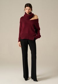 Donna che indossa un maglione a maglia bordeaux off-shoulder, pantaloni neri su misura e stivali con punta a stiletto neri, in piedi su uno sfondo uniforme.