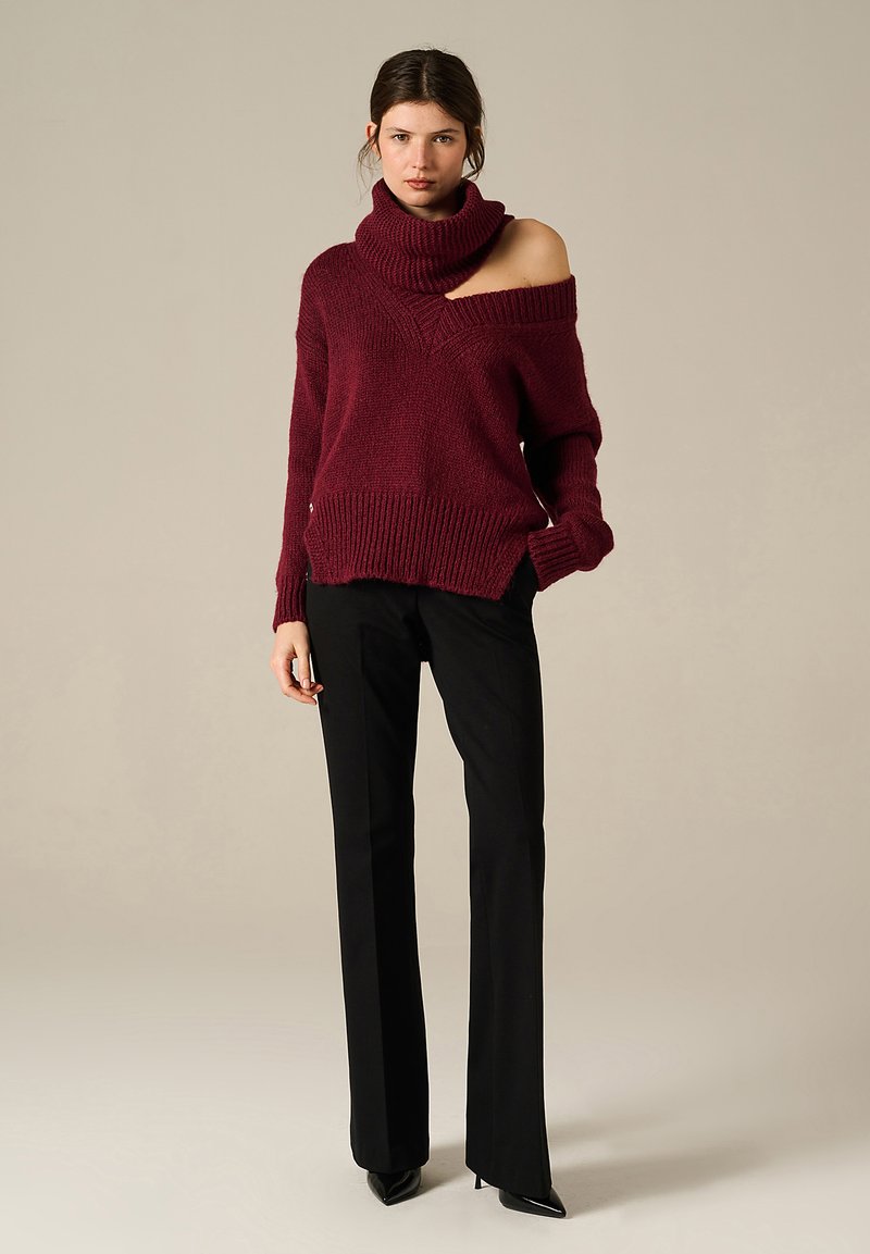 Donna che indossa un maglione a maglia bordeaux off-shoulder, pantaloni neri su misura e stivali con punta a stiletto neri, in piedi su uno sfondo uniforme.