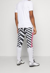 Man i vit träningströja och sportbyxor med svart-vitt zick-zackmönster, röda sidostripes och vita fotbollsskor, stående med ryggen mot kameran.