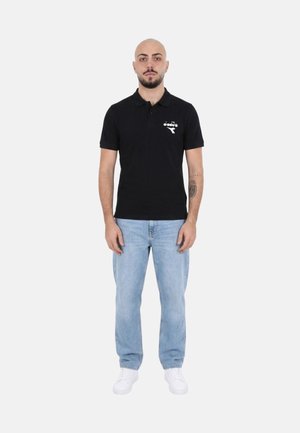 Uomo in piedi che indossa una polo nera con logo bianco, jeans azzurri, sneakers bianche e mostra un tatuaggio sull'avambraccio.