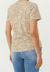 Top de manga corta con un patrón floral en tonos beige, blanco y negro. Tejido suave con cuello redondo y un corte relajado, combinado con jeans azul claro.