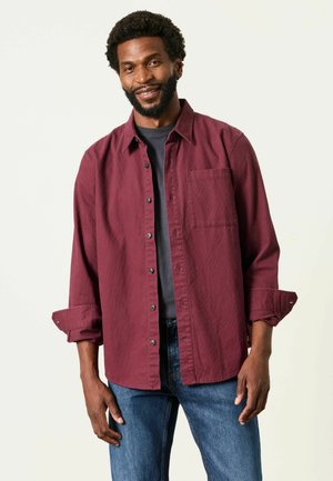Lächelnder Mann mit Bart, der ein bordeauxrotes Hemd über einem dunklen T-Shirt und blauen Jeans trägt und vor einem hellen Hintergrund steht.