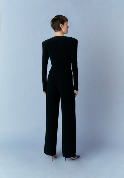 Black long-sleeve jumpsuit with a fitted bodice and wide legs, featuring gathered texture at the back. Worn with blue high-heel shoes.
