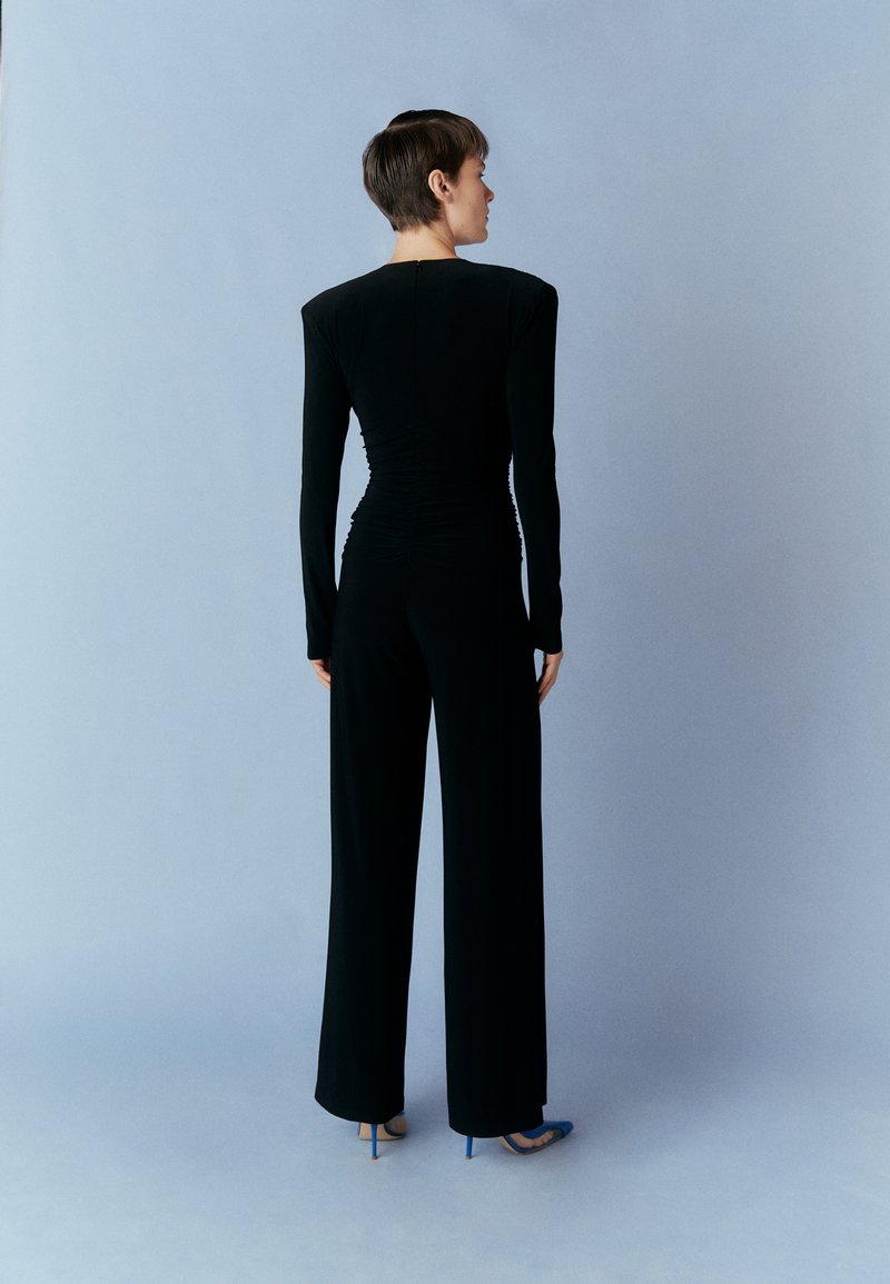 Zwarte jumpsuit met lange mouwen, een getailleerd lijfje en wijde pijpen, met geplooide textuur aan de achterkant. Draagt bijpassende blauwe hoge hakken.