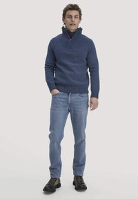 Dunkelblauer Strickpullover mit hohem Kragen und Reißverschluss, kombiniert mit hellblauen Jeans und dunklen Schnürstiefeln, stehend vor einem neutralen Hintergrund.