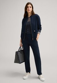 Marineblau bomberjacke mit zwei vorderen Taschen, kombiniert mit einem gemusterten Oberteil und einer taillierten Hose. Weiße Sneaker und eine marineblaue gemusterte Tote-Tasche runden den Look ab.