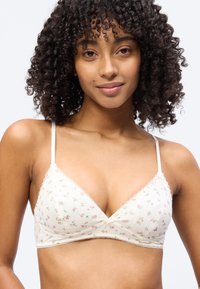 Νεαρή γυναίκα με σγουρά μαλλιά που φοράει ένα λευκό, φλοράλ σχέδιο bralette με λεπτές τιράντες, χαμογελώντας απαλά πάνω σε απλό φόντο.