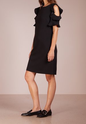 Vrouw draagt een zwarte knielange jurk met gerimpelde cold-shoulder mouwen en zwarte instap-loafers met gespsdetail.