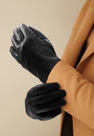 Black leather gloves with a smooth texture, fitted design, and four seams on the back, worn on a wrist, paired with a tan coat.