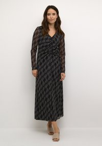 Dolga črna obleka s prosojnimi dolgimi rokavi, ki ima ponavljajoč se geometrijski vzorec v beli barvi. Obleka z drapirano zasnovo in nabranim pasom.