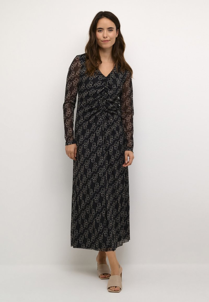 Dolga črna obleka s prosojnimi dolgimi rokavi, ki ima ponavljajoč se geometrijski vzorec v beli barvi. Obleka z drapirano zasnovo in nabranim pasom.