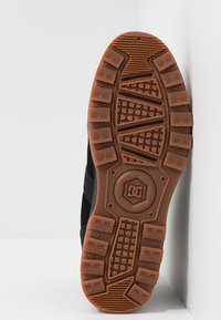 Czarna podeszwa zewnętrzna buta sportowego wykonana z teksturowanej gumy. Posiada wzorzystą przyczepność oraz okrągłe logo, kontrastujące z brązowym bieżnikiem.