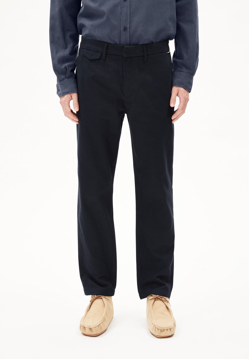 Zwarte katoenen broeken met een rechte snit, voorzien van een tailleband met riemlussen en zijzakken, gecombineerd met lichtbeige moccasin-stijl schoenen.