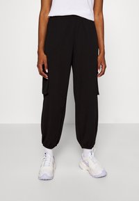 Pantalon cargo noir avec une ceinture élastique, des poches latérales et des poignets ajustés. Porté avec des chaussures de sport blanches avec des accents violets.