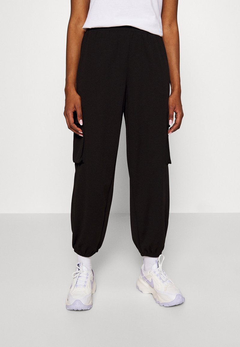 Pantalon cargo noir avec une ceinture élastique, des poches latérales et des poignets ajustés. Porté avec des chaussures de sport blanches avec des accents violets.