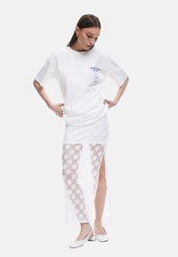Vit bomull t-shirt med grafiskt detalj, kombinerad med en lång vit spetskjol med blommönster och sidslits, bärs med vita ballerinas.