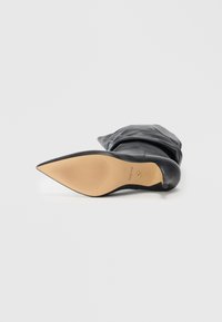 Black pointed-toe ankle boot with a smooth leather upper, a soft fold-over design, and a tan rubber sole featuring linear tread patterns.