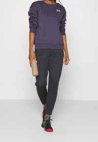 Sudadera azul marina con puños y dobladillo de punto, combinada con pantalones jogger negros. La persona sostiene una botella de agua beige y lleva zapatillas negras con detalles rojos.