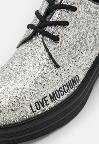 Glittrande sneakers i silver med svarta snören, med en svart strukturerad sula och texten "LOVE MOSCHINO" i fet stil.