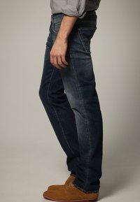 Jeans de mezclilla en azul oscuro, estilo de pierna recta con un ligero desgaste. Presenta cinco bolsillos y un sutil detalle de costura. Combinados con zapatos marrones.