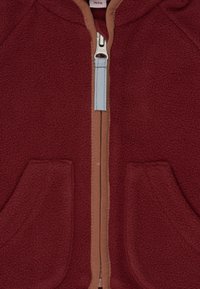 Rote Fleecejacke mit einem hellbraunen Reißverschluss und zwei Vorderfächern. Der Reißverschluss hat einen hellblauen Zipper. Weicher Stoff insgesamt.