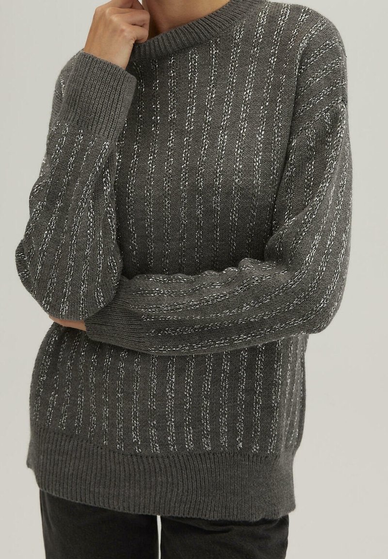Pull en tricot gris avec une texture côtelée et des rayures verticales métalliques argentées. Il présente un col rond et des manches ajustées.