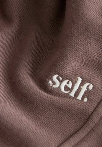 Ruskea kangas, jossa on pehmeä pinta, ja valkoinen brodeerattu logo, jossa lukee "self." sijoitettuna näkyvästi alaosaan.