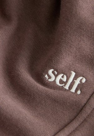 Barna textil, puha tapintással, fehér hímzett logóval, amelyen az olvasható: "self." A logó a alsó szakaszban hangsúlyosan helyezkedik el.