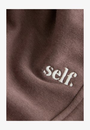 Barna textil, puha tapintással, fehér hímzett logóval, amelyen az olvasható: "self." A logó a alsó szakaszban hangsúlyosan helyezkedik el.