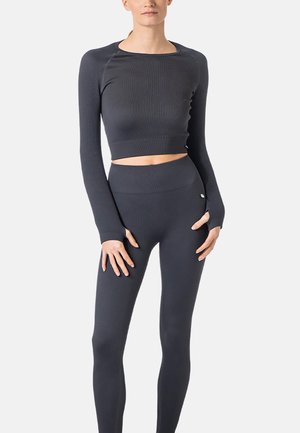 Frau trägt dunkelgraues, geripptes, langärmeliges Crop-Top und passende hoch taillierte Leggings vor schlichtem Hintergrund.