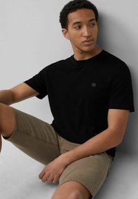 Schwarzes Baumwoll-T-Shirt mit kurzen Ärmeln und Rundhalsausschnitt. Kleines Logo-Detail auf der Brust. Kombiniert mit beigen Shorts. Glatte Textur.