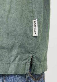 Grøn linnedskjorte med et mærke, der står "ORIGINALS." Kanten har sideslids, og den er parret med denimjeans i en blå vask.