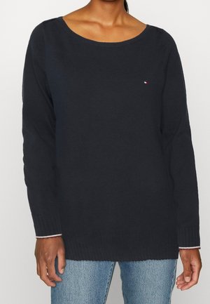 Frau trägt einen schlichten dunkelblauen Pullover mit langen Ärmeln und kleinem Logo auf der Brust sowie hellblaue Jeans, steht vor einem weißen Hintergrund.