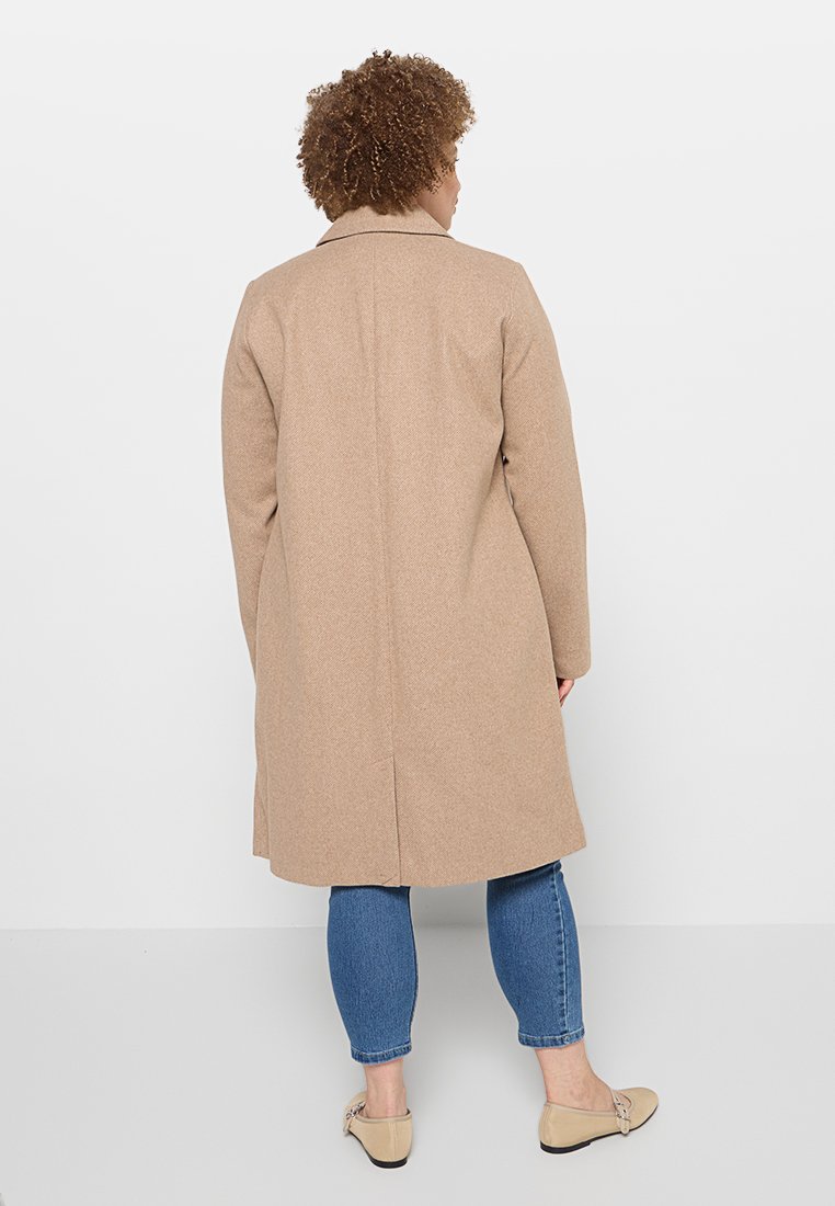 Beige Wollmischungsjacke mit Reverskragen und Rückenbelüftung, kombiniert mit blauen Jeans und hellen Slippern. Glatte Textur.