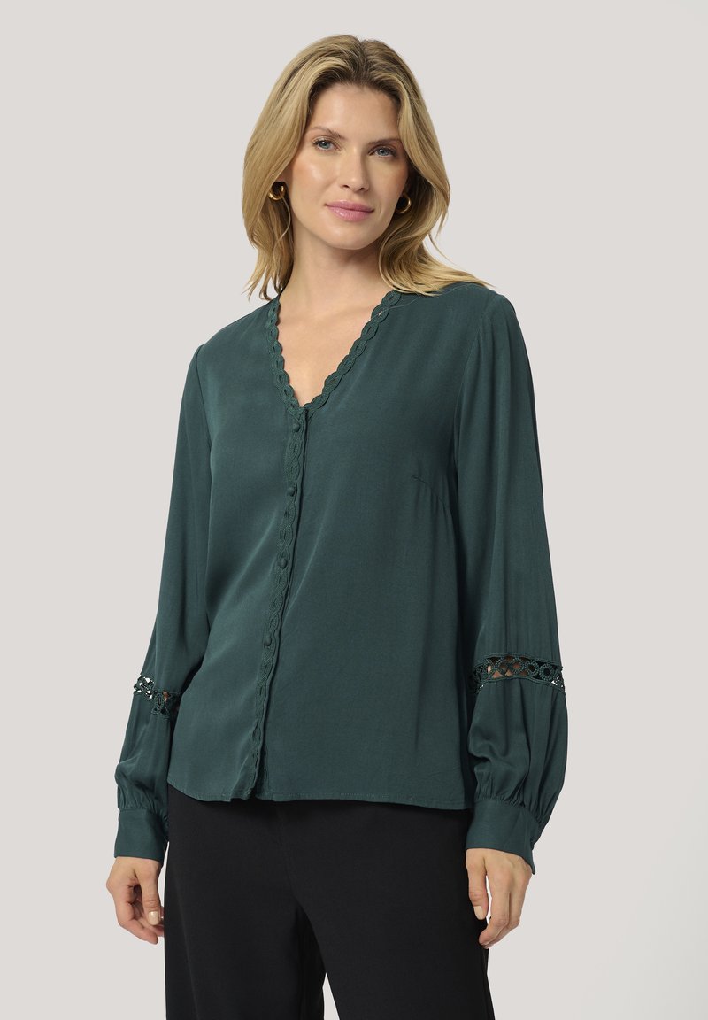 Blusa verde scuro con scollo a V, bordo ondulato e maniche lunghe a sbuffo con dettagli in pizzo ai polsini. Tessuto liscio e leggero.