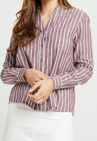 Chemise à manches longues boutonnée mauve avec des rayures verticales blanches. Dispose d'un col en V, d'une poche avant et d'un ourlet légèrement arrondi.