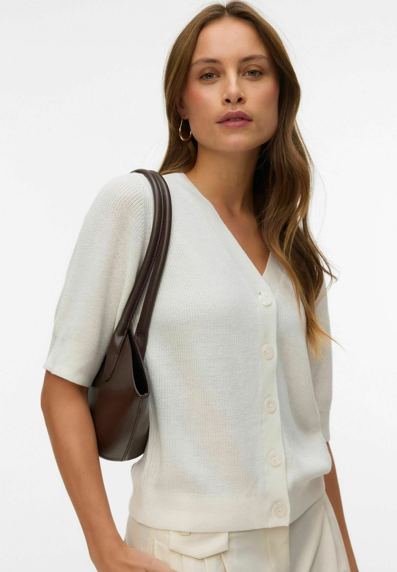 Femme portant un cardigan blanc à manches courtes boutonné, un pantalon crème, des boucles d'oreilles créoles dorées et un sac à bandoulière marron foncé.