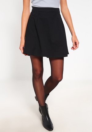 Kvinde iført sort A-formet nederdel, gennemsigtige sorte strømpebukser, sorte ankelstøvler og en lysegrå ærmeløs top på hvid baggrund.