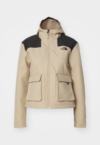Chaqueta con capucha beige y detalles en negro, cuenta con un cierre de cremallera en el frente, dos grandes bolsillos frontales y un logo en la parte superior izquierda. Textura de tela suave.